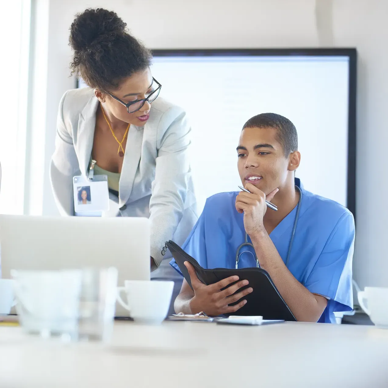 Healthcare professional in scrubs reviewing documents while collaborating with a colleague in a business suit in a meeting room.